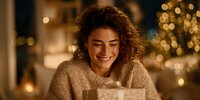 Young woman with curly hair smiling while opening gift box at home with festive bokeh lights in background, creating warm holiday atmosphere.