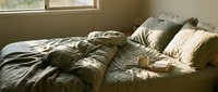 Unmade bed with olive green and cream bedding in warm sunlight streaming through window, conveying relaxation and comfort for bedroom design projects.