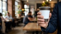Person holding blank white coffee cup with brown lid in modern cafe environment with blurred customers and warm lighting in background.