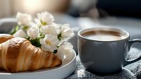 Fresh buttery croissant served on white plate with delicate white flowers and hot cappuccino in gray ceramic cup on cozy textured surface in soft morning light.