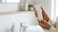 Person with medium skin tone holding blank white cosmetic bottle in modern bathroom setting, ideal for skincare, beauty product marketing, and wellness branding.