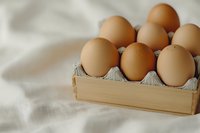 Fresh brown eggs arranged in wooden box with cardboard divider on white textile background, soft natural lighting creates gentle shadows and depth.