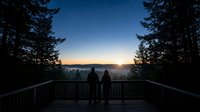 Silhouetted figures stand on wooden deck overlooking misty valley at sunrise, framed by evergreen forest trees and golden sky. Perfect for travel, nature, and lifestyle concepts.