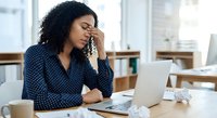 Stressed African American businesswoman with headache working on laptop in modern office, touching forehead while experiencing work pressure and fatigue.