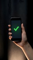 Hand holding smartphone displaying bright green checkmark on dark screen against black textured background. Success, approval, confirmation, and validation concept for mobile apps, websites, and digital services.