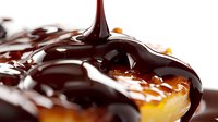 Liquid dark chocolate sauce pouring onto golden caramel surface creating glossy swirls and drips in macro photography against white background.