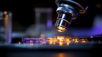 Microscope objective lens examining illuminated microchip circuit board with glowing orange and purple lights in dark laboratory setting, scientific research concept.