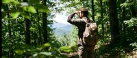 Young adult male with medium skin tone wearing brown outdoor gear, holding binoculars while birdwatching in dense forest. Lush green foliage surrounds him with mountain vista visible in background.