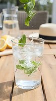Fresh mint leaves dropping into sparkling water glass on wooden table with lemon slices creating refreshing summer drink with bubbles and splash.