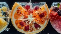 Fresh citrus orange slice with raspberries frozen in clear ice cube with water drops and bubbles, macro photography showing detailed texture and cold effect.