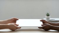 Two people of different ethnicities holding a blank white tablet device between them at a table with a small potted succulent plant, suitable for app mockups and digital product presentations.