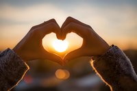 Hands forming heart shape silhouette against golden sunset sky with warm glowing light shining through fingers creating romantic love symbol for Valentine's Day.