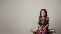 Young girl with braided hair and dark skin tone wearing burgundy traditional clothing, sitting on stacked money against neutral background for financial education or wealth concept.