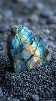 Raw labradorite crystal displaying vibrant blue green orange iridescent flash on dark volcanic sand beach natural mineral specimen geology collection.