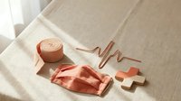 Coral medical face mask, bandage roll, heartbeat line and cross symbol arranged on linen surface, healthcare and wellness concept.
