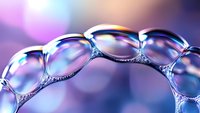 Macro shot of soap bubbles forming delicate chain pattern with purple and blue bokeh background, showing iridescent surface and light reflections.