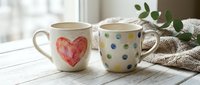 Ceramic mugs with heart and polka dot patterns sit on white wooden table near window with natural light and cozy textile decor.