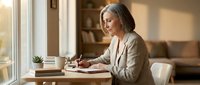 Mature woman with gray hair and light skin tone, wearing a beige cardigan, sits at a wooden desk by a window, writing in a notebook with a coffee cup and books nearby, suitable for lifestyle, wellness, and productivity content.