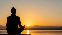 Person meditating in lotus position silhouetted against golden sunrise over calm lake water creating peaceful mindfulness moment for wellness and relaxation.