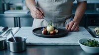 Chef plating gourmet dish with vegetables and sauce on black plate in professional kitchen. Male cook garnishing fine dining food preparation.