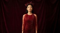 Woman wearing ornate crown and red beaded gown stands against burgundy velvet backdrop. Asian ethnicity, regal appearance, luxury fashion and costume design.