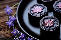 Dark chocolate pralines with delicate pink flower design on black ceramic plate, decorated with fresh purple spring flowers on wooden table background.