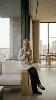 Young blonde businesswoman enjoying coffee break in modern office lounge with city skyline view, relaxing on contemporary furniture during workday.