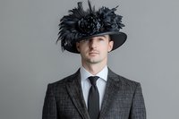 Elegant Caucasian man in gray suit and black tie wearing extravagant wide-brim hat with feathers, studio portrait on gray background.