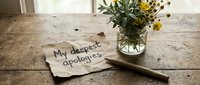 Handwritten my deepest apologies note on crumpled brown paper beside a pencil and wildflowers in a glass jar on a rustic wooden table.