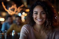 Young African American woman with curly hair smiling warmly at evening restaurant setting, wearing casual pink sweater against blurred bokeh lights background.