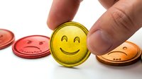 Smiling yellow token held between fingers with red and copper coins blurred in background, isolated on white for customer satisfaction, feedback and positive rating concepts.