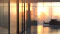 Atmospheric sunset view through glass office partition, creating soft bokeh effect with silhouettes of modern workspace furniture and warm golden light.