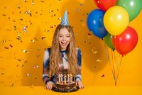 Young woman celebrating birthday with chocolate cake and colorful balloons on orange background with confetti falling around her.