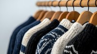Cozy knitted sweaters in navy blue, white and black hanging on wooden hangers in closet, showing texture and pattern details of winter wardrobe collection.