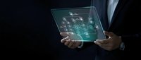 Businessman holding transparent digital tablet displaying futuristic interface with glowing icons and network connections on dark background for technology presentations.