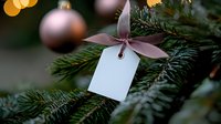 Blank white gift tag with pink ribbon hanging on Christmas tree branch with ornaments and bokeh lights in background, festive holiday mockup design space.