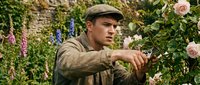 Young male gardener in work clothes and cap tending flowering plants and roses in lush garden setting, suitable for gardening, horticulture, and lifestyle content.