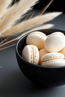 White macarons in black ceramic bowl with pampas grass decoration on dark background. Elegant French pastry close-up with soft natural lighting.