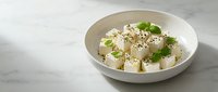 Fresh white cheese cubes with herbs and sesame seeds in ceramic bowl on marble surface for healthy Mediterranean cuisine and cooking.