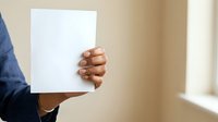 Man holding blank white paper sheet, close-up of hand and arm in casual blue sleeve against neutral beige wall background for mockup and branding purposes.