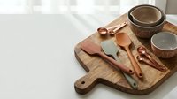 Rustic wooden cutting board with copper kitchen utensils and ceramic bowls arranged for cooking preparation on white marble countertop surface.