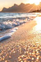 Golden sunlight sparkles on wet sand as waves roll onto shore during sunset, creating magical bokeh effect against mountain silhouettes in background.
