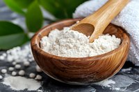 Wooden bowl filled with white baking flour and bamboo spoon on dark background with green leaves and white towel, close up view for cooking concept.