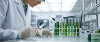 Male scientist in white lab coat examining plant samples with magnifying glass in modern laboratory with green test tubes for biotechnology research.