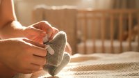 Woman hands holding tiny knitted baby socks in warm sunlight near wooden crib preparing for newborn arrival and motherhood journey.