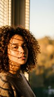 Young mixed-race woman with curly hair enjoying warm sunlight through window blinds creating shadow patterns on face in cozy indoor setting.
