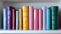 Row of colorful hardcover books standing on white shelf, spines in pastel shades of blue, purple, pink, yellow and green creating minimalist library composition.