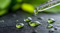 Glass dropper releasing clear liquid drop onto dark surface with green-tinted water droplets scattered around, creating natural lens effect against blurred botanical background.