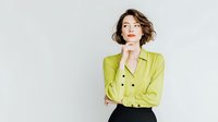 Young woman with short brown hair in lime green blazer posing thoughtfully against white background, hand on chin in contemplative gesture.