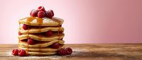 Stack of golden pancakes with fresh raspberries and syrup on wooden table against pink background for breakfast menu design and food photography.